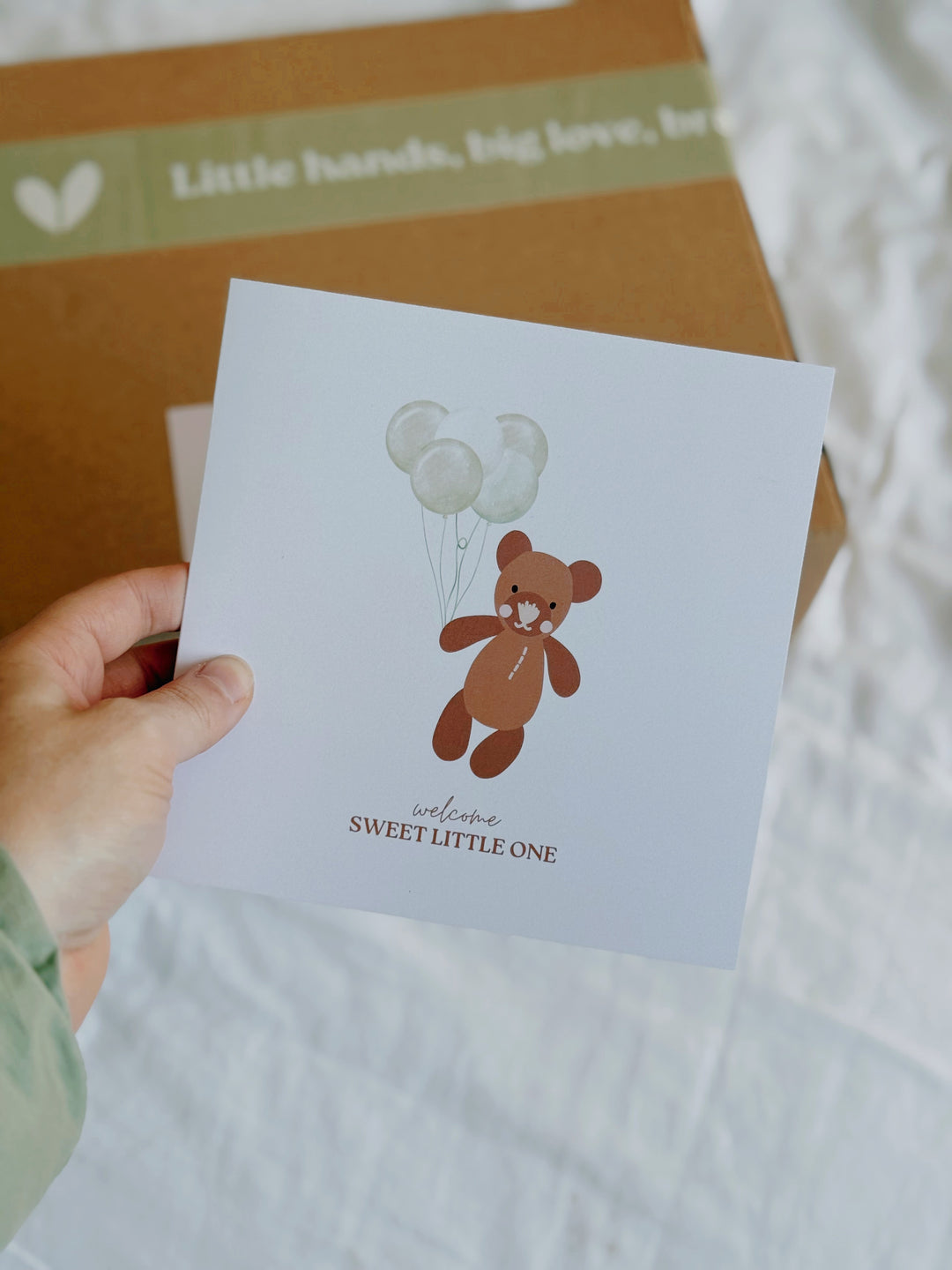 Hand holding a card with a bear and balloons illustration and text 'Welcome Sweet Little One'.